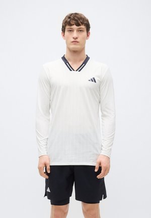 Fehér, hosszú ujjú sportpóló, függőleges csíkokkal, V-nyakkivágással és tengerészkék díszítéssel, valamint az Adidas logóval a bal mellrészen. Fekete rövidnadrággal viselve.