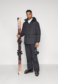 Homem com jaqueta e calças de esqui pretas segura esquis e óculos de esqui pretos contra um fundo simples.