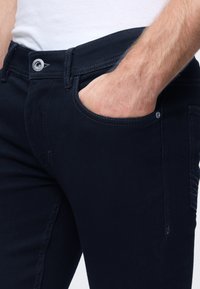 Tmavomodré deninové nohavice so slim fit strihom, s gradientovou textúrou, prednou vreckom a kontrastnými švami. Viditeľný gombík a pútka na opasok.