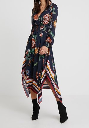 Kobieta ubrana w granatową, długorękawową sukienkę w kwiaty z asymetrycznym dołem i paskiem w paski, w zestawie z czarnymi botkami za kostkę.