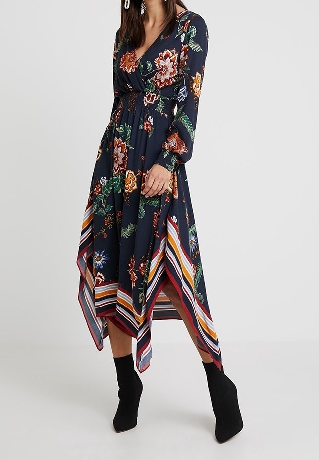 Femme portant une robe longue à manches longues bleu marine à fleurs avec un ourlet en forme de mouchoir et une bordure rayée, associée à des bottines noires.