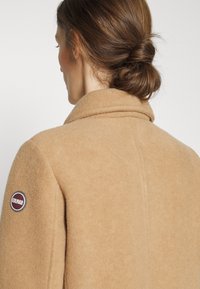 Hellbrauner Wollmantel mit weicher Textur, ausgestattet mit einem hohen Kragen und einem runden Logo-Patch am linken Ärmel. Rückansicht zeigt klare Linien.