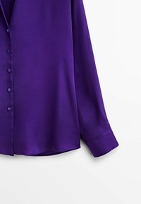 Chemise en satin violet à boutons avec manches longues, présentant une texture lisse et un éclat subtil, agrémentée de boutons espacés uniformément.