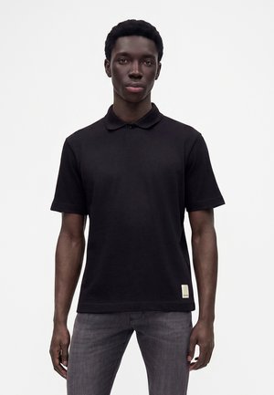 Ung mand med mørk hud iført en sort kortærmet polo-shirt og grå jeans, stående mod en ensfarvet hvid baggrund.