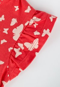 Tissu rouge avec un bord froncé présentant un motif imprimé de papillons blancs, détail en gros plan de la couture et de la texture du vêtement.