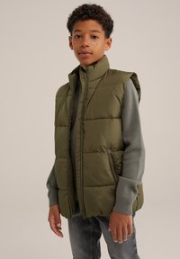 Olivgrön puffväst med hög krage, dragkedja och två sidofickor, bärs över en grå långärmad skjorta med en kviltad design.