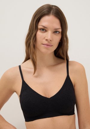 Vrouw met lichte huid en golvend bruin haar, gekleed in een zwarte kanten bralette, tegen een effen lichte achtergrond, vooruitkijkend.