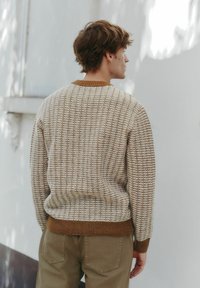 Beige knit sweater with a textured pattern, brown ribbed trim at the collar and hem, paired with light khaki pants.