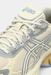 Asics futócipő világos bézs hálóból, szürke részletekkel és kék díszítéssel. Fókuszban a fényvisszaverő csíkok, sík fűzők és párnázott talp.