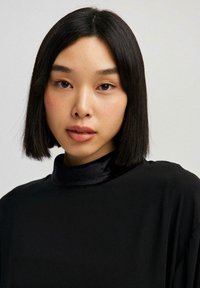 Sort top med en elegant, høj halsudskæring og glat tekstur. Håret er glat, skulderlangt og rammer ansigtet ind. Naturlig makeup.