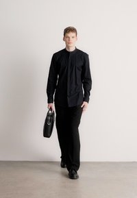 Fekete hosszú ujjú ing magas nyakkivágással, fekete nadrággal és fekete cipővel párosítva. A modell egy kis fekete táskát tart, amelyen fém díszítés található.