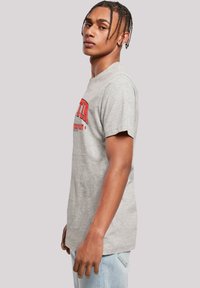 Light gray cotton t-shirt with short sleeves and a crew neck. Features red text graphic on the front. Model poses sideways.