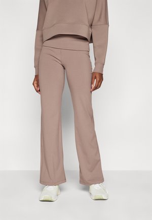 Person iført beige vidde bukser og matchende sweatshirt, stående foran en ensfarvet lys baggrund med synlige hvide sneakers.