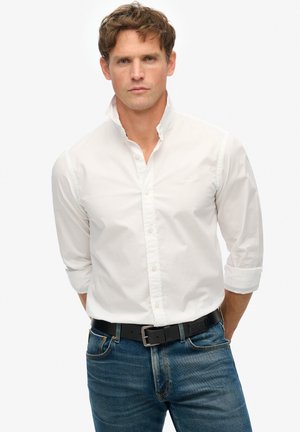 Witte button-up shirt met een kraag, opgestroopte mouwen, gecombineerd met blauwe spijkerbroeken en een zwarte riem. De stof lijkt glad, met subtiele stiksels.