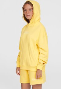 Felpa gialla con cappuccio e pantaloni corti abbinati, realizzati in materiale morbido. La felpa presenta una grande tasca frontale e il logo "Surf Society" in bianco.