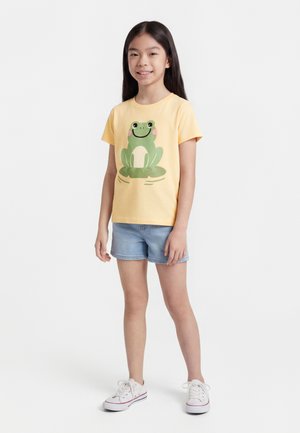 Tyttö seisoo ja hymyilee, pukeutuneena keltaiseen t-paitaan, jossa on vihreä sammakkoaihe, vaaleansiniset farkkushortsit ja valkoiset lenkkarit.