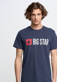 Jūras zila kokvilnas T-krekls ar īsām piedurknēm, kuram ir balts "BIG STAR" logo un sarkans zvaigznes akcents kvadrātā. Standarta piegriezums.