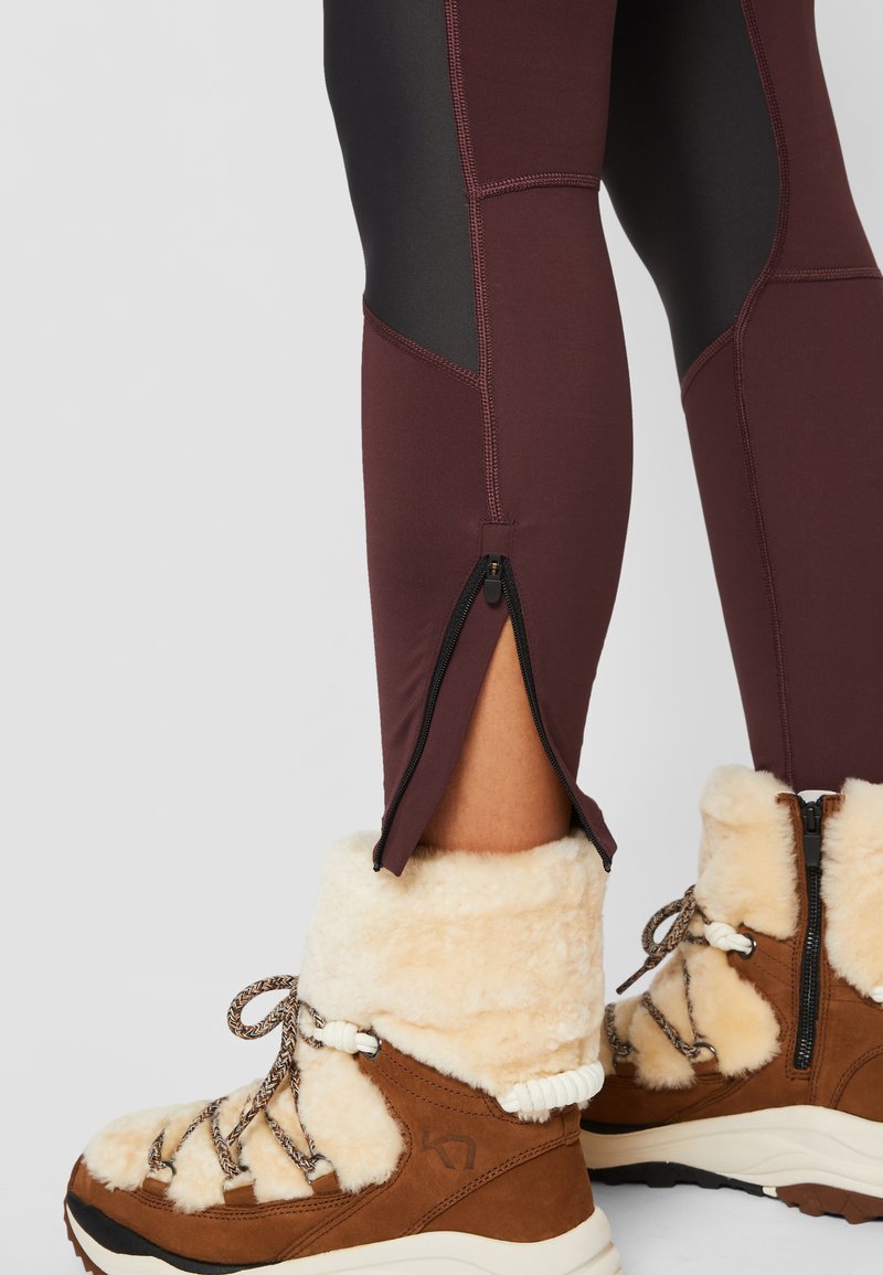 Bordeauxfarbene Leggings mit schwarzen Einsätzen, ausgestattet mit einem Seitenreißverschluss am Knöchel. Getragen mit braunen, mit Fell gefütterten Winterstiefeln.