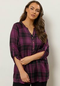 Violetti ja musta ruudullinen paita, jossa on V-pääntie, käärittävät hihat ja napituksessa yksityiskohtia. Pehmeä kangas, joka istuu löyhästi ja siinä on hillitty rakenne.