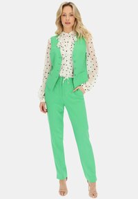 Zielona kamizelka z guzikami, biała bluzka w białe kropki z falbanami, dopasowane zielone spodnie, przezroczysta tekstura i jasnobeżowe szpilki.