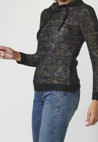 Pletený černý svetr s vícebarevnými flíčky, kapucí a stahovacími šňůrkami; doplněný modrými džínami s květinovou výšivkou po straně.