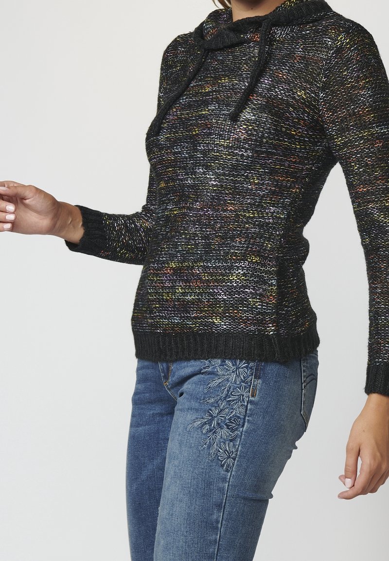 Pletený černý svetr s vícebarevnými flíčky, kapucí a stahovacími šňůrkami; doplněný modrými džínami s květinovou výšivkou po straně.