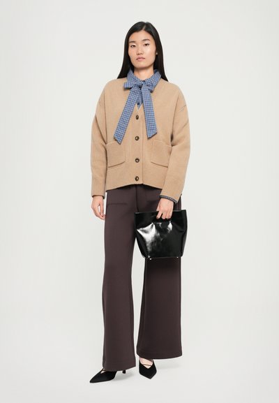 Γυναίκα στέκεται φορώντας ένα μπεζ cardigan πάνω από μπλε καρό μπλούζα με φιόγκο, φαρδείς σκούρους καφέ παντελόνες, μαύρες γόβες, και κρατά μια μικρή μαύρη τσάντα.