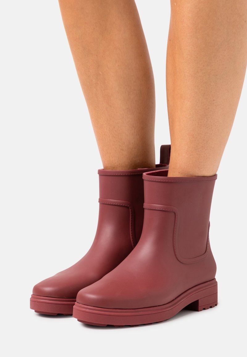 Paire de bottes en caoutchouc bordeaux mates mi-mollet portées sur des jambes nues, debout sur une surface lisse de couleur gris clair.