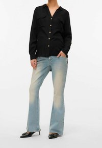 Mujer vestida con una camisa negra de botones, vaqueros acampanados de color azul claro desgastados y tacones negros de puntera afilada con un detalle de lazo.