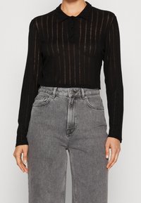 Czarna polo z długim rękawem w pionowy wzór, z kołnierzykiem i zapięciem na trzy guziki. W połączeniu z jasnoszarymi spodniami z wysokim stanem.