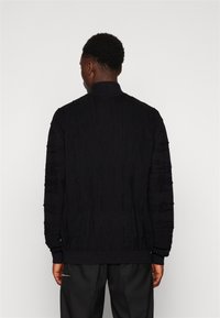 Black knitted sweater featuring a textured pattern with vertical lines, ribbed collar, and cuffs. Back view with long sleeves.
