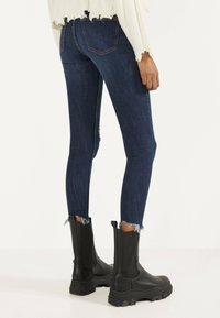 Jeans ajustados de color azul oscuro con dobladillos deshilachados, combinados con botines negros gruesos y un suéter blanco desgastado.