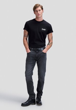 Unge mand med tatoveringer iført en sort Lee t-shirt, mørke jeans, sort bælte og sorte støvler, stående med hænderne i lommerne.