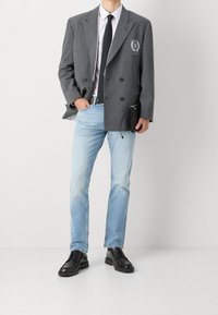 Šedý kostkovaný dvouřadý blejzr, bílá košile, černá kravata, světle modré džínové kalhoty a černé šněrovací boty. Drží černou kapsu.