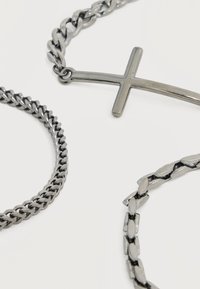 Trois chaînes en métal argenté, dont une avec un pendentif en forme de croix latérale, exposées sur une surface blanche.