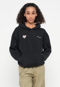 Zwarte hoodie met een voorvak, voorzien van een roze hartontwerp en "PEGA"-tekst. De stof lijkt zacht en heeft een relaxed fit met een trekkoordcapuchon.