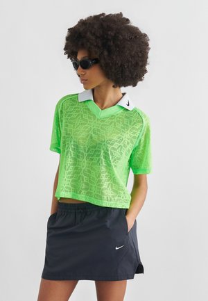 Femeie cu păr creț purtând ochelari de soare negri, tricou polo cu mâneci scurte și model verde neon și fustă neagră Nike, cu mâinile în buzunare.
