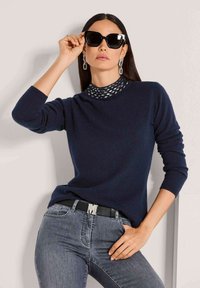 Frau mit langen dunklen Haaren trägt schwarze Sonnenbrille, silberne Kettenohrringe, navy-langärmliges Oberteil und graue Jeans mit schwarzem Gürtel und posiert vor einer Wand.