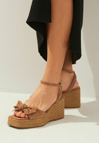 Braune Wildleder-Wedge-Sandalen mit einer gewebten Jute-Plattform, ausgestattet mit einem großen Schleifendetail an der Zehe und einem Knöchelriemen mit einer verstellbaren Schnalle.