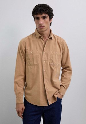 Camisa de manga larga color canela hecha de algodón, con dos bolsillos en el pecho, cuello de botón y un corte relajado. Se lleva con unos jeans de mezclilla oscuros.