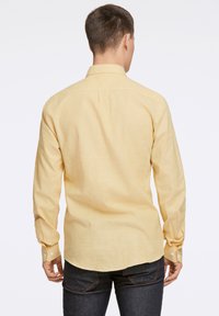 Lange mouwen, lichtgele shirt met een kraag, enkele knopen bij de manchetten en een licht afgeronde zoom, gecombineerd met donkere jeans.