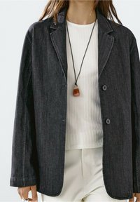 Blazer gris foncé avec un col cranté, deux boutons et des poches latérales, associé à une chemise blanche côtelée et un collier pendentif.