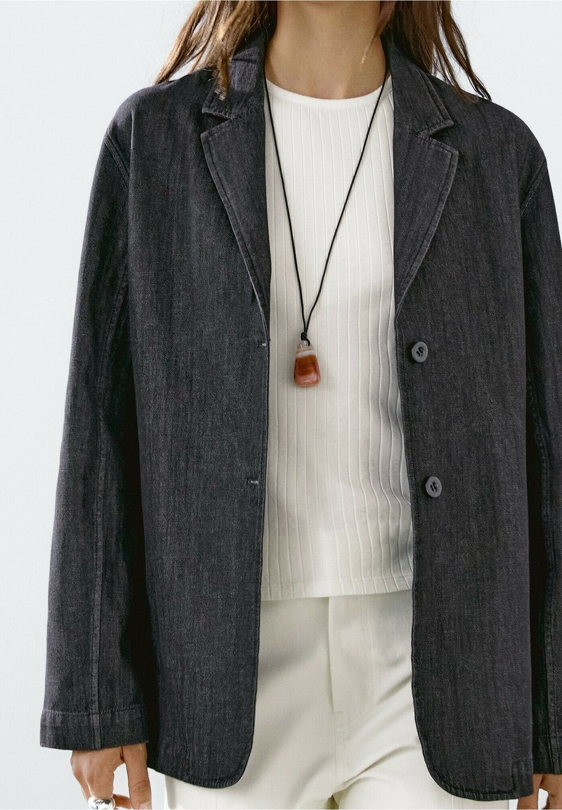 Blazer gris foncé avec un col cranté, deux boutons et des poches latérales, associé à une chemise blanche côtelée et un collier pendentif.