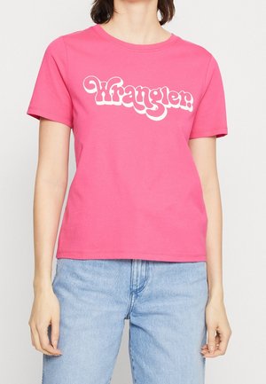 Vrouw draagt felroze Wrangler T-shirt en lichtblauwe spijkerbroek, staand tegen een effen lichte achtergrond.