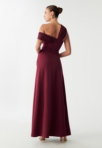 Bordo večernja haljina s jednom perlicanom naramenicom, uskim korzetom i lepršavom suknjom. Glatka tekstura tkanine i elegantna silueta.