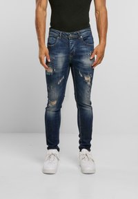 Tmavě modré skinny džíny s poškozeným vzhledem, lehkým vyblednutím a několika roztrhnutími, doplněné o bílé tenisky.