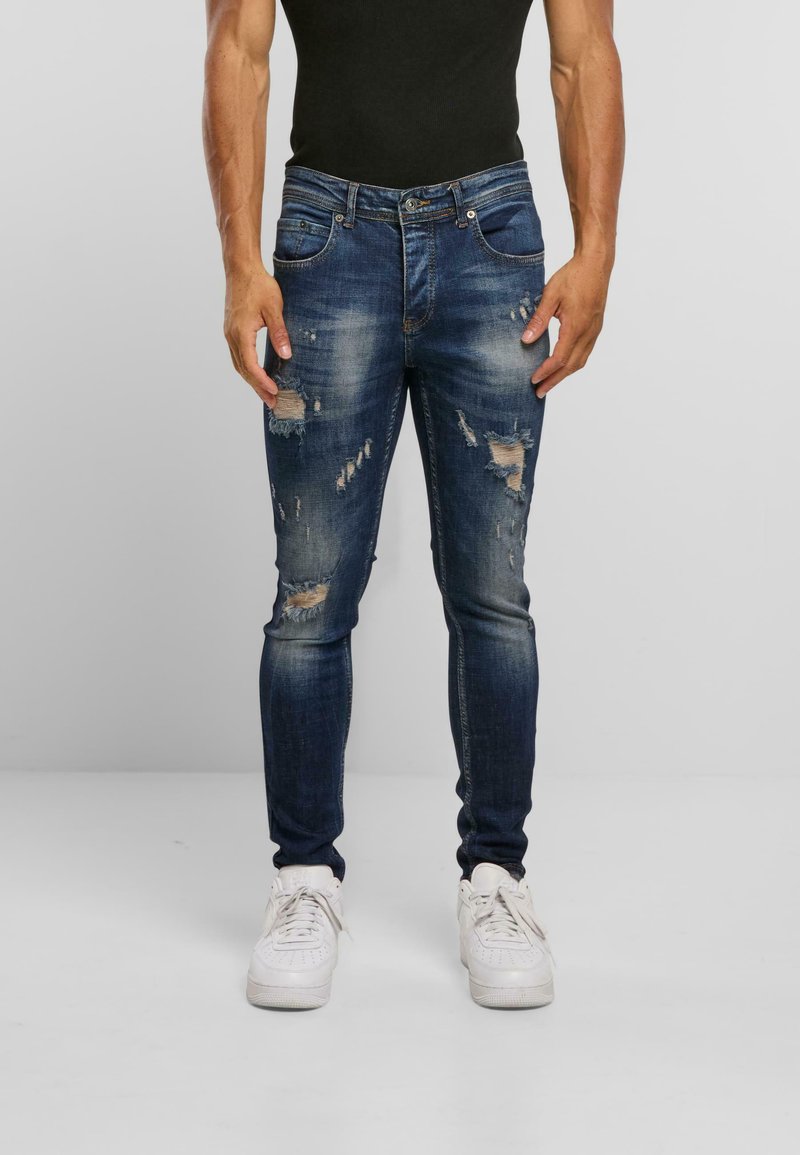 Tmavě modré skinny džíny s poškozeným vzhledem, lehkým vyblednutím a několika roztrhnutími, doplněné o bílé tenisky.