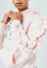 Sudadera con capucha de color rosa claro con detalles de volantes en las mangas, puños acanalados y cordones con texto. La tela parece suave y gruesa, lo que sugiere calidez.