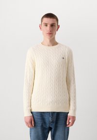 Cream cable-knit sweater with a round neckline, ribbed cuffs, and hem, paired with blue jeans. Logo detail on left chest.