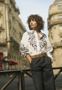 White button-up shirt featuring black floral embroidery and puffed sleeves, paired with dark denim pants and a brown belt with a decorative buckle.